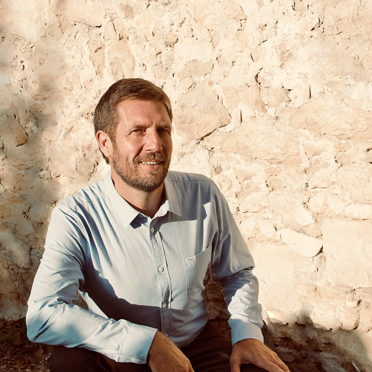 Portrait homme souriant en chemise bleue Lagoon, mur pierre Homme souriant, barbe, chemise bleue Lagoon, assis contre mur en pierre ancien sous lumière du soleil couchant.