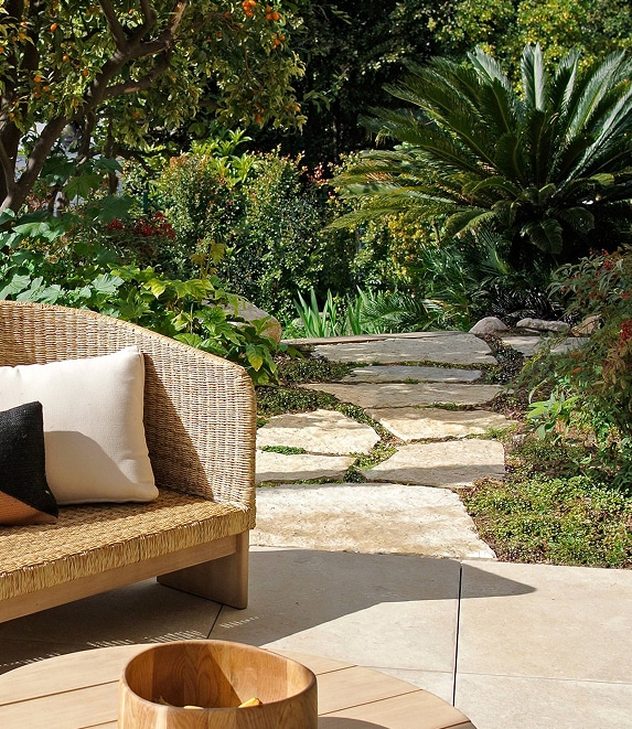 Jardin luxuriant : chemin pierre et salon extérieur Canapé en osier sur terrasse pavée menant à un chemin de pierres dans un jardin luxuriant avec oranger et palmier.