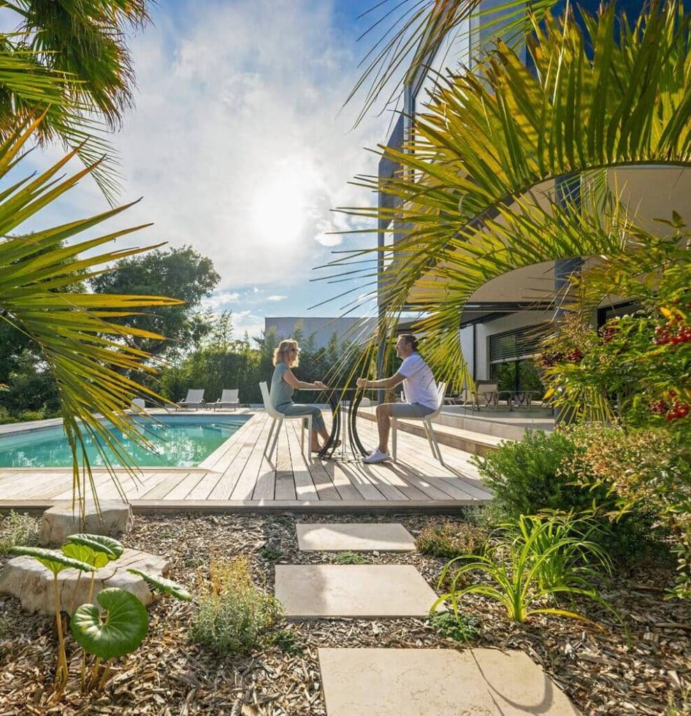 Luxe et détente: Piscine, terrasse en bois et jardin tropical Couple souriant sur une terrasse en bois au bord d'une piscine, conversant. Jardin tropical avec palmiers et maison moderne sous le soleil.