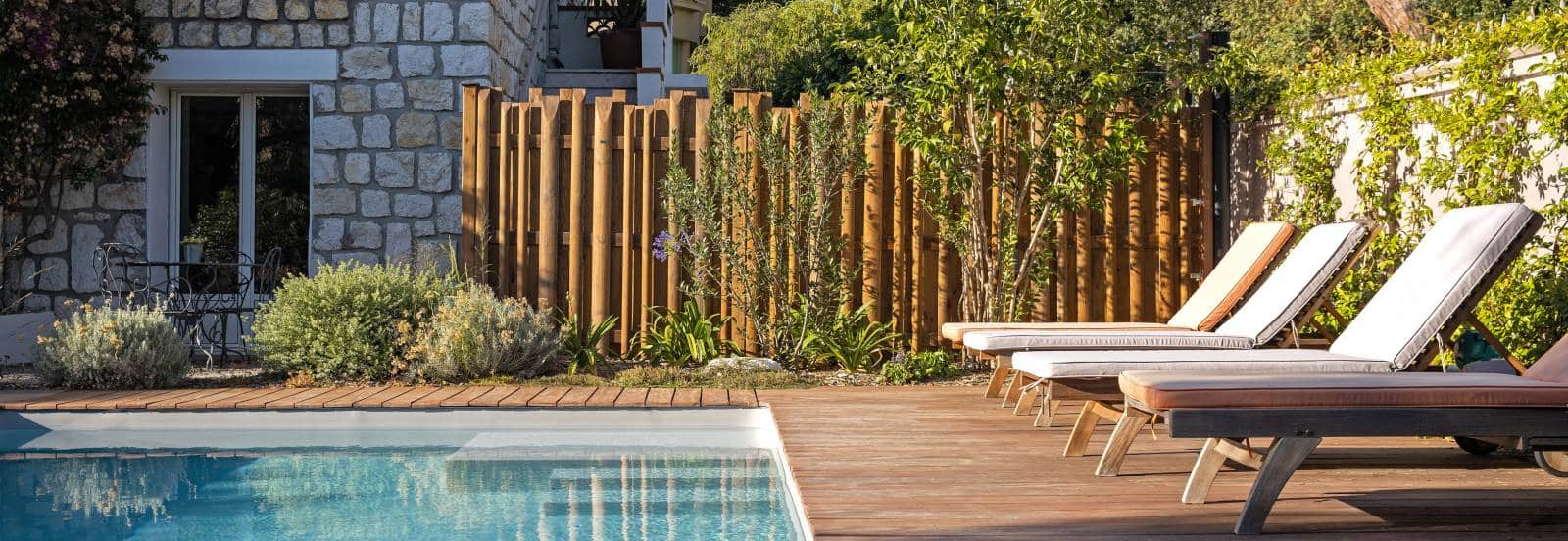 Bordure de piscine en bois avec transats et clôture en rondins de bois, près d'un mur de pierre.
