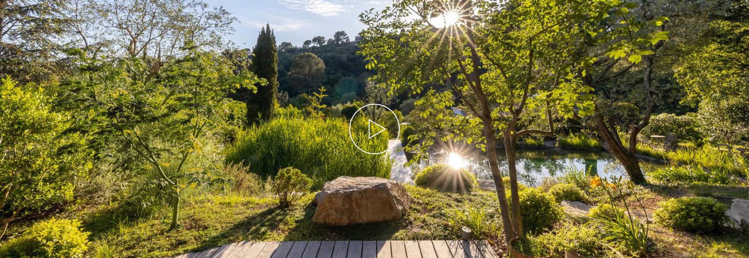 Jardin luxuriant baigné de soleil avec un plan d'eau, un rocher au premier plan et une icône de lecture vidéo au centre.